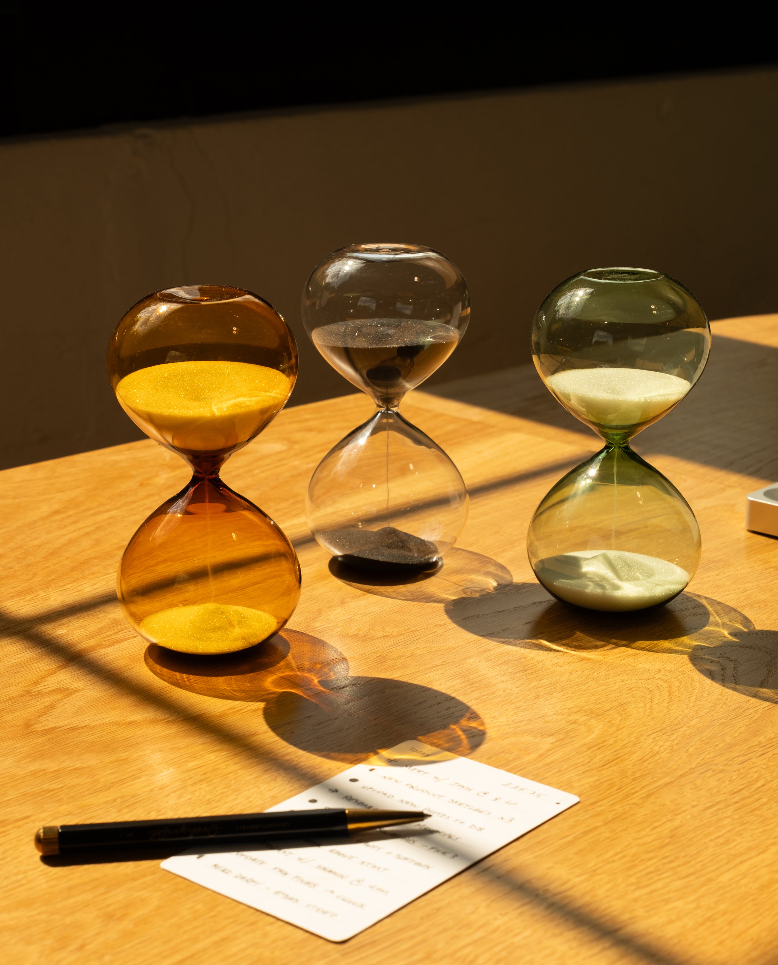 A 15 Minute Hourglass with green sand sits on a wooden table, sunlight casting shadows across the surface and highlighting the analog timer. A pen and a small notepad with writing are also on the table.