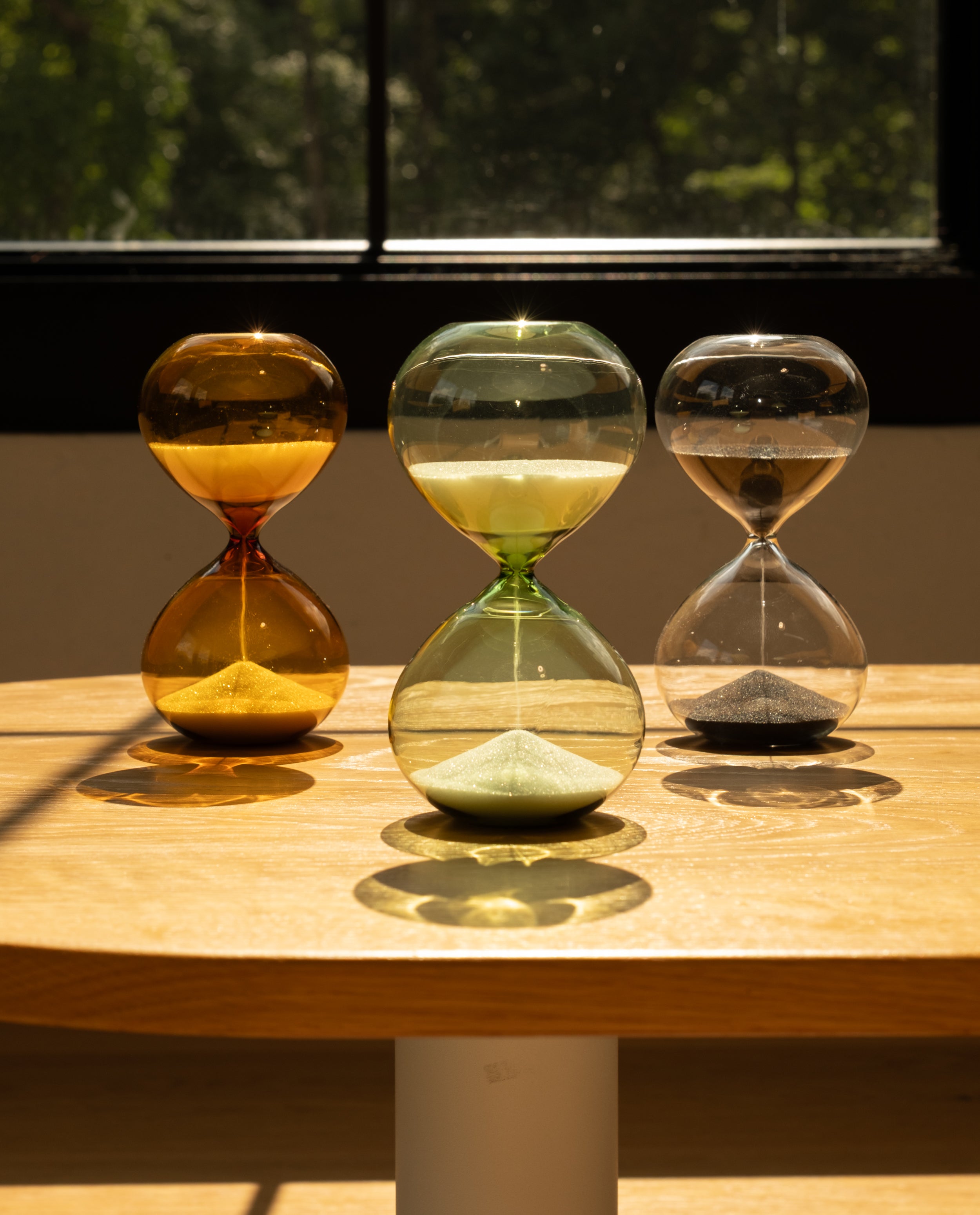 A single 15 Minute Hourglass with green sand serves as a visual focus tool on a sunlit wooden table; trees and sunlight are visible outside the window in the background.