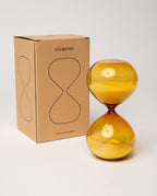A 15 Minute Hourglass (Amber) filled with fine yellow glass sand sits next to a brown cardboard box labeled UGMONK, featuring a simple hourglass graphic on the front. This elegant analog timer is a practical focus tool, ideal for timing tasks or meditation. The background is plain white.