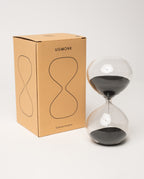 A 15 Minute Hourglass (Clear) with black sand, featuring a sleek analog design, is placed beside a brown box labeled UGMONK that displays a minimalist hourglass graphic and the text “15 Minute Hourglass” on the front.