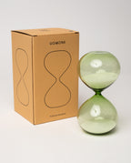 A 15 Minute Hourglass (Green) with white sand and a green glass body rests next to a brown box labeled UGMONK 15 Minute Hourglass, which features an hourglass outline printed on the front. The analog timer, serving as a visual representation of time, is displayed against a plain white background.