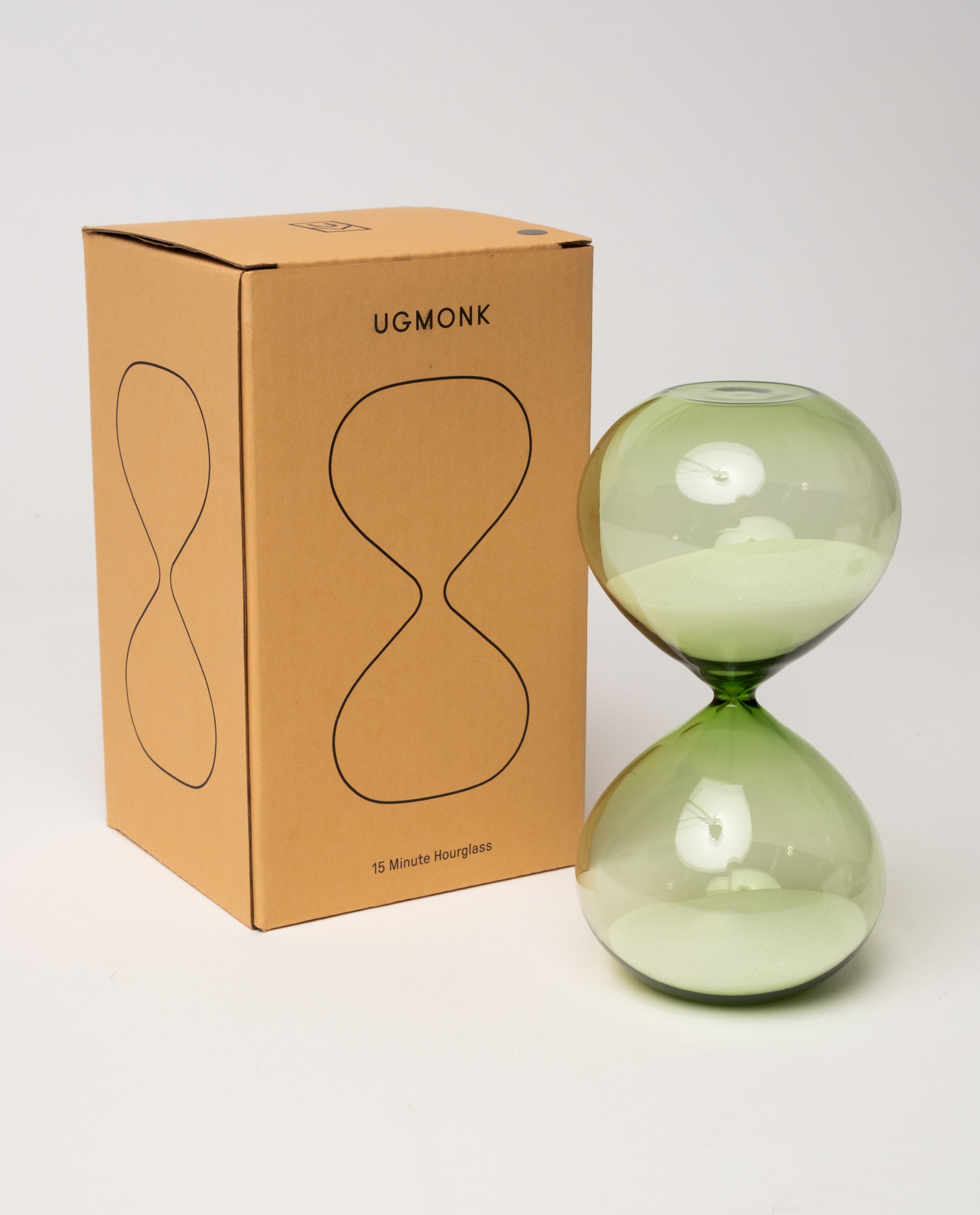 A 15 Minute Hourglass (Green) with white sand and a green glass body rests next to a brown box labeled UGMONK 15 Minute Hourglass, which features an hourglass outline printed on the front. The analog timer, serving as a visual representation of time, is displayed against a plain white background.