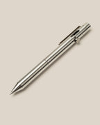 A sleek, metallic silver INVENTERY Bolt Action Pen (Stainless) with a clip and push-button mechanism is set diagonally from bottom left to top right, shown against a plain light beige background.