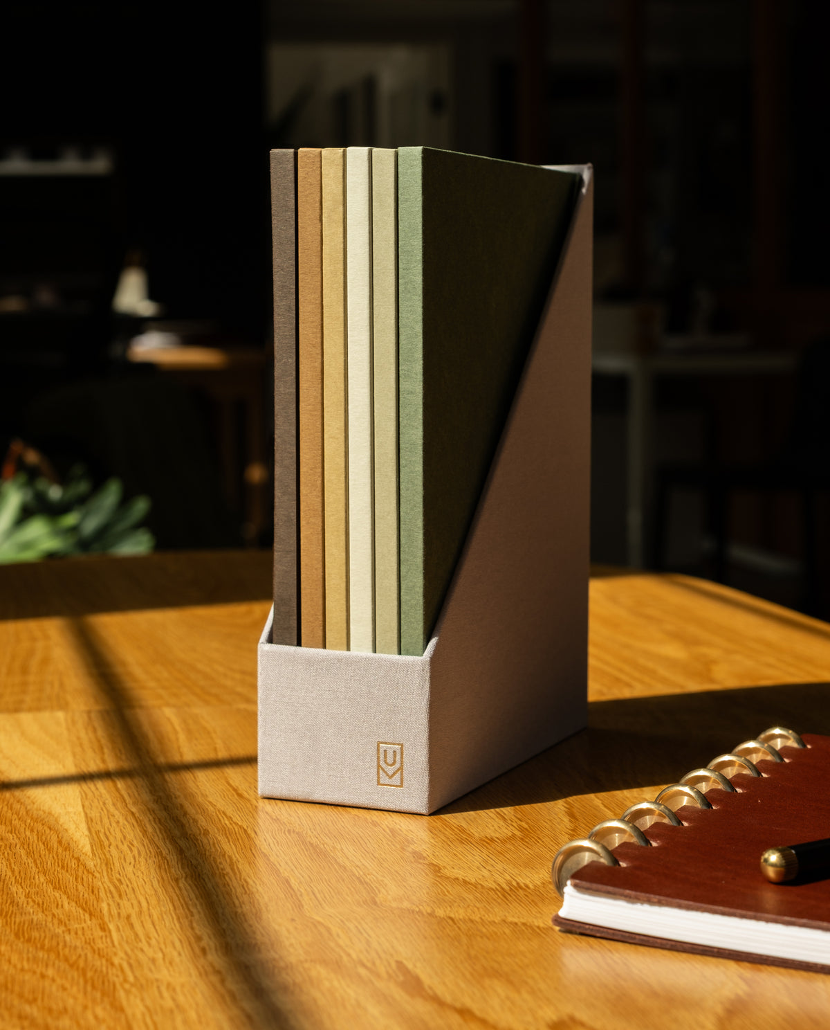 Six refillable Discbound Journal Archives in neutral colors stand upright in a fabric holder on a wooden desk, creating an elegant storage solution, while a spiral-bound notebook sits nearby. Sunlight casts shadows across the surface.