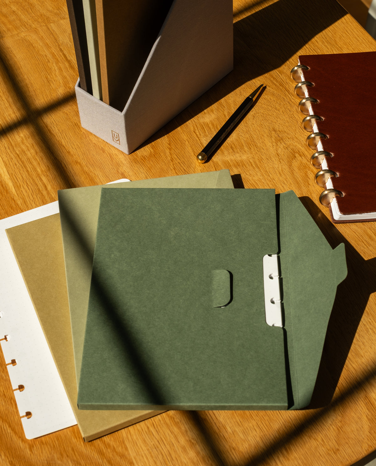 Green, beige, and brown folders rest on a wooden desk alongside a brown notebook, a black pen, and the Discbound Journal Archive—a vertical box organizer made specifically for journal storage. Sunlight casts gentle shadows across the workspace, emphasizing this versatile setup—perfect for keeping your Discbound Journal Archive within easy reach.
