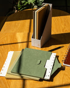 On a wooden table, the Discbound Heirloom Journal Bundle (Brown Leather 6″ x 8.5″) rests alongside a green notebook and a pen. In the background, a gray vertical file holder displays additional notebooks, while sunlight casts soft shadows across the scene.