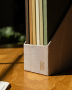 A Discbound Journal Bundle (Standard 6″x 8.5″ Journal), fabric-covered and featuring a small geometric logo on the front, stands on a sunlit wooden desk. The bundle’s design evokes the tactile appeal of a refillable notebook and contains several colored folders inside, while some greenery is softly blurred in the background.