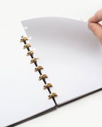 A hand lifts a blank, dotted page in the Warehouse Item - Discbound Heirloom Journal (Black/Brass), featuring brass rings, against a white background.