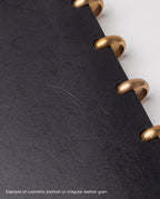 Close-up of a black full grain leather cover with brass rings and a faint scratch in the center, showing a cosmetic blemish or irregular leather grain—an ideal find for our warehouse sale on this Warehouse Item - Discbound Heirloom Journal (Black/Brass).