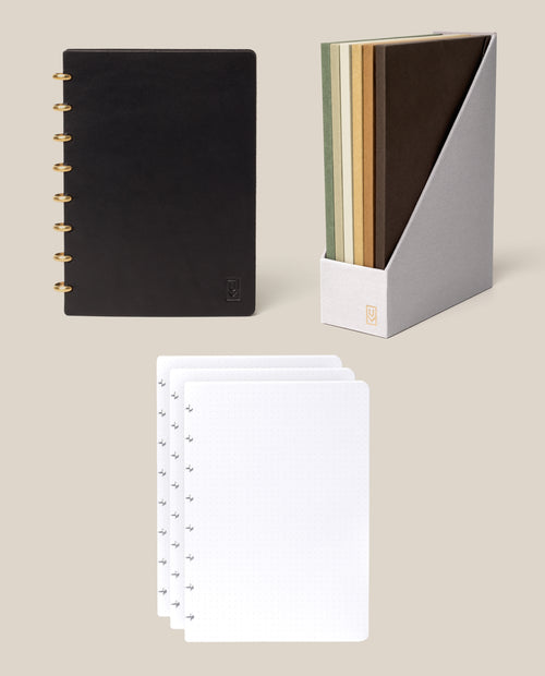 A Discbound Heirloom Journal Bundle in black leather (Standard 6″ x 8.5″) is displayed with four loose dot grid paper pages and a gray holder containing five full-grain leather journals in assorted earth tones, all set against a beige background.