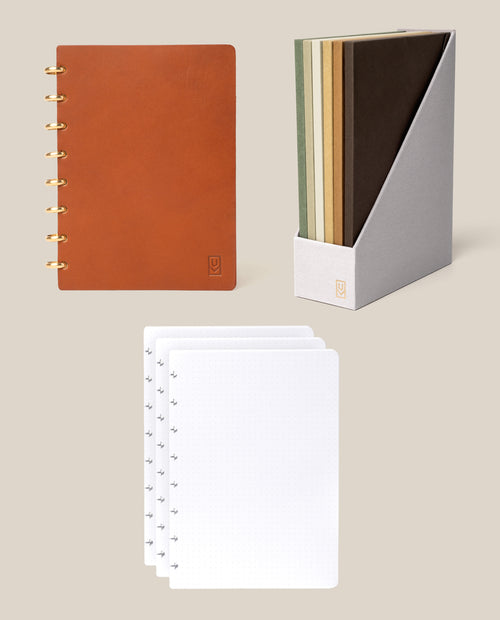 Displayed on a beige background is the Discbound Heirloom Journal Bundle in Tan Leather (Standard 6″ x 8.5″), featuring a brown spiral notebook with dot grid pages, four blank white dotted sheets, and four assorted-color notebooks arranged in a gray holder.