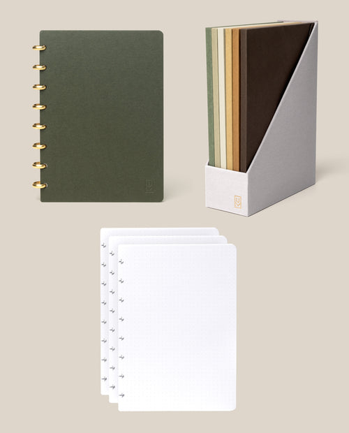 The Discbound Journal Bundle (Standard 6″x 8.5″ Journal), including a green ring-bound refillable notebook, a gray holder containing four notebooks in green, tan, brown, and mustard yellow, and three sheets of dot grid paper, is arranged on a beige background.