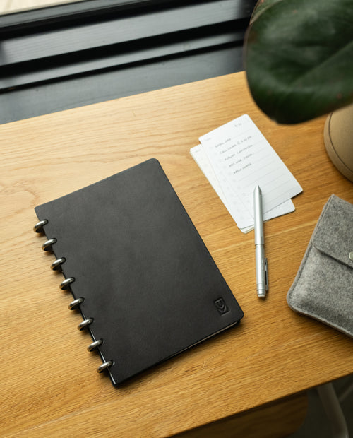 A Discbound Heirloom Journal + 3 Refills (Black Leather - Standard 6″ x 8.5″), accompanied by a silver pen, small notepaper, and a gray felt pouch, rests on a wooden desk near a window with a potted plant partially visible.