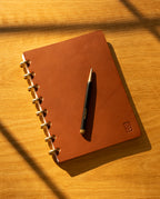 The Discbound Heirloom Journal in tan leather (Standard 6" x 8.5") rests on a wooden surface with diagonal shadows, gold rings glinting and a black and gold pen placed neatly on top.
