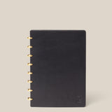 The Discbound Heirloom Journal (Black Leather - Standard 6" x 8.5") with gold rings is centered on a beige background, featuring a small embossed logo in the lower right corner of its black leather cover.