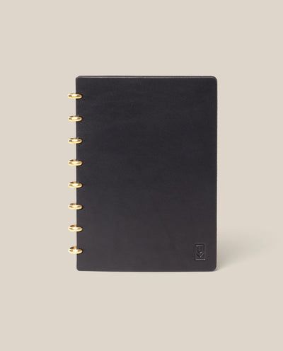 The Discbound Heirloom Journal (Black Leather - Standard 6" x 8.5") with gold rings is centered on a beige background, featuring a small embossed logo in the lower right corner of its black leather cover.