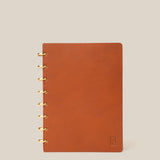The Discbound Heirloom Journal (Tan Leather - Standard 6" x 8.5") stands upright on a plain beige background, featuring gold ring binding on the left and a small embossed logo in the bottom right corner of its refillable leather cover.