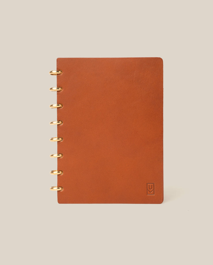 The Discbound Heirloom Journal (Tan Leather - Standard 6" x 8.5") stands upright on a plain beige background, featuring gold ring binding on the left and a small embossed logo in the bottom right corner of its refillable leather cover.