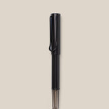 A black LAMY AL-star Rollerball pen with a transparent section near the tip is vertically centered on a plain beige background, highlighting its smooth writing experience and refillable rollerball design.