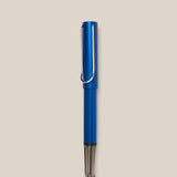 A LAMY AL-star Rollerball (Oceanblue) with a metal clip stands vertically on a light beige background, featuring smooth writing and a stylish design.