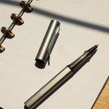 A graphite LAMY AL-star Rollerball with its cap off rests on an open, dotted notebook, demonstrating smooth writing in sunlight that casts shadows across the wooden surface.
