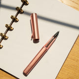 A LAMY AL-star Rollerball (Flamingo), cap off, rests on an open dotted spiral notebook atop a wooden surface, its anodized aluminum body gleaming in natural sunlight.