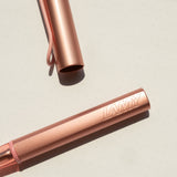 A close-up of the LAMY AL-star Rollerball (Flamingo) with its cap removed shows its anodized aluminum body on a light dotted surface, and "LAMY" embossed on the barrel.