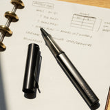 A LAMY AL-star Rollerball (Black) with its cap off rests on an open dotted notebook showing handwritten project notes. Sunlight casts shadows across the page, highlighting the pen’s smooth writing experience.