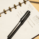 A LAMY AL-star Rollerball (Black) rests on an open dotted notebook filled with handwritten notes and a diagram, set atop a light wooden surface. Sunlight casts shadows, emphasizing the pen's smooth writing experience.