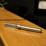 A graphite LAMY AL-star Rollerball lies diagonally on a sunlit wooden surface, with natural light casting shadows and highlighting its metallic finish, offering smooth writing performance.