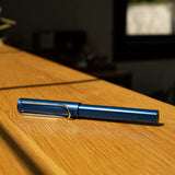 A LAMY AL-star Rollerball (Oceanblue) sits on a wooden surface in natural sunlight, its sleek design highlighted against a softly blurred background with a window and indoor decor.