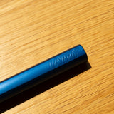 A close-up of a blue LAMY AL-star Rollerball (Oceanblue), the refillable pen known for smooth writing, embossed with the brand name and resting diagonally on a wooden surface in bright sunlight.