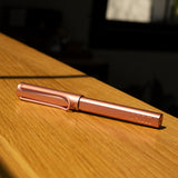The LAMY AL-star Rollerball (Flamingo), featuring a metallic pink anodized aluminum body, lies on a wooden surface in sunlight, casting gentle shadows and highlighting its modern design.