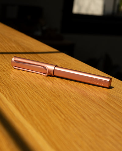 The LAMY AL-star Rollerball (Flamingo), featuring a metallic pink anodized aluminum body, lies on a wooden surface in sunlight, casting gentle shadows and highlighting its modern design.