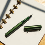 A pine green LAMY AL-star Rollerball with its cap off rests on an open dotted notebook with gold rings, placed on a sunlit wooden surface—perfect for smooth writing.