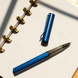 A LAMY AL-star Rollerball (Oceanblue) with its cap off rests on an open dotted notebook with gold rings, set on a wooden surface in bright sunlight, highlighting its smooth writing and refillable rollerball design.