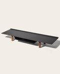 The Gather Large Monitor Stand (Black/Walnut) features a minimalist powder-coated steel surface in flat black with four short walnut legs, designed as a sleek, space-saving desk organizer on a light background.