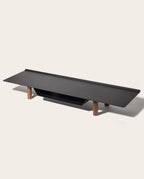The Gather Large Monitor Stand (Black/Walnut) features a minimalist powder-coated steel surface in flat black with four short walnut legs, designed as a sleek, space-saving desk organizer on a light background.