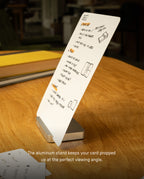 A note card with handwritten notes is propped up on a silver aluminum stand from the Analog Starter Kit - BOGO Special, resting on a wooden table. The softly blurred background adds focus to the scene. Text at the bottom reads: The aluminum stand from the Analog Starter Kit keeps your task cards propped up at the perfect viewing angle—a simple solution for an effective paper card system.
