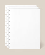Three sheets of the Large Discbound Journal Refill - Dot Grid (8.5″ x 11″)—white and perforated—are stacked with offset edges on a beige background, making them ideal for expanding your discbound notebook.