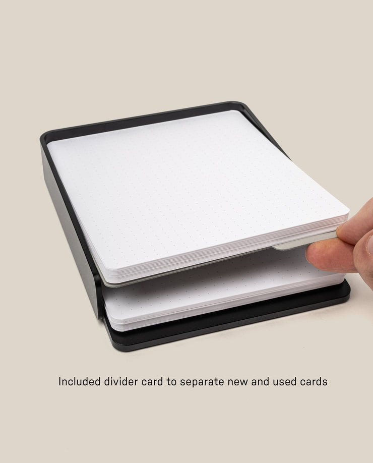A hand places a divider card between two stacks of large dot grid cards in a black tray. Text: The Analog Capture Kit - Large (Black Aluminum) includes a divider to separate new and used cards—perfect for organizing your workflow.