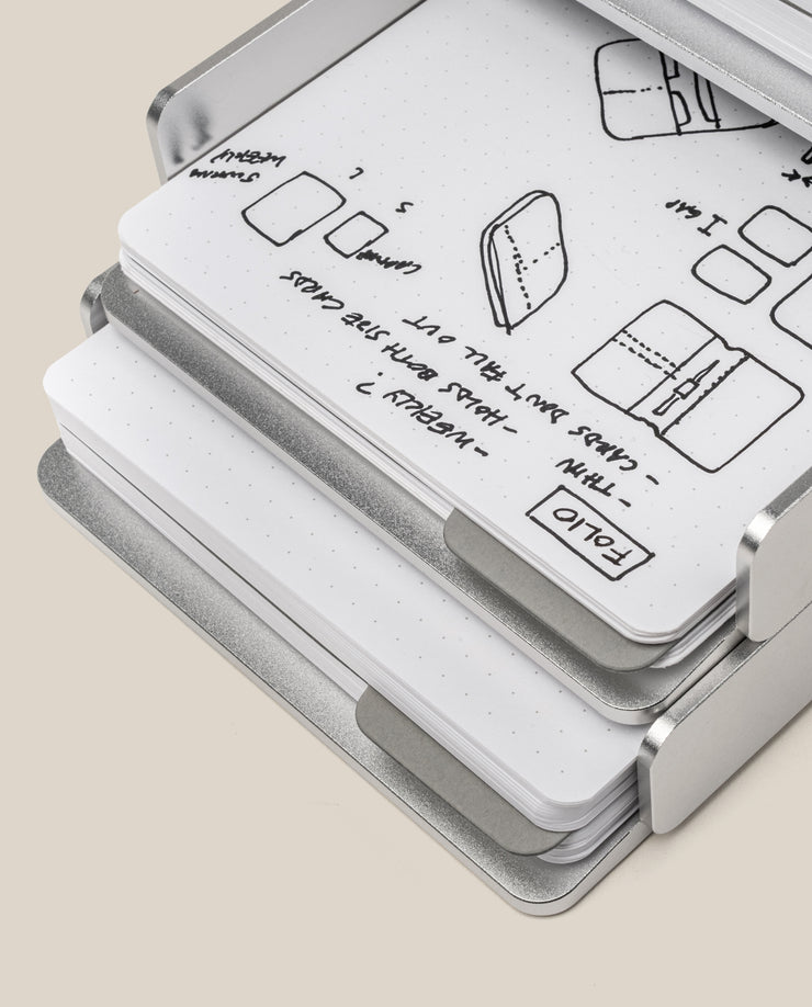 The Analog Capture Kit - 2 Large (Silver Aluminum) is displayed open, revealing dot grid notebooks and cards with black ink sketches and notes on folding and wallet designs on crisp white pages.