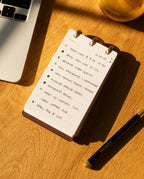 Notebook with to-do list on a wooden desk with a laptop and pen.