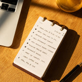 Notebook with to-do list on a wooden desk with a laptop and pen.