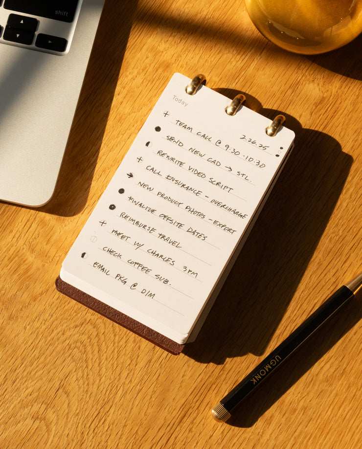 Notebook with to-do list on a wooden desk with a laptop and pen.