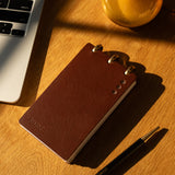 Brown notebook and pen on a wooden surface with a laptop and glass in the background