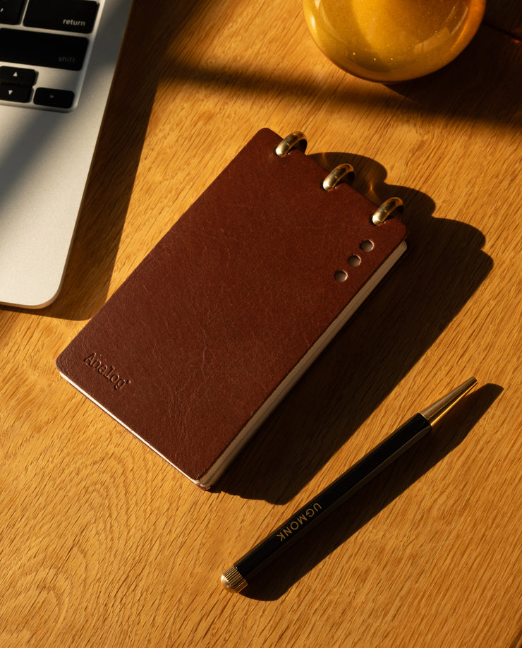 Brown notebook and pen on a wooden surface with a laptop and glass in the background