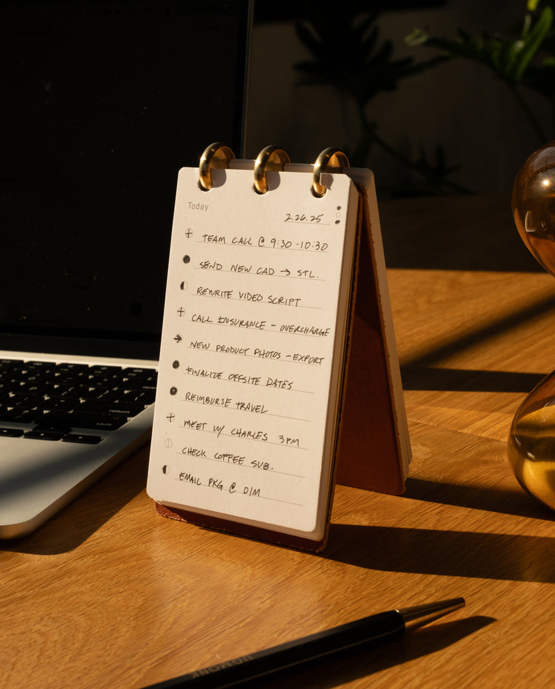 A Leather Discbound Analog + 3 Refills (Tan) rests on the desk beside a laptop and an hourglass, as sunlight casts shadows across the wooden surface. Inside, to-do lists with tasks like team call, script rewrite, and emailing merge classic elegance with everyday productivity.