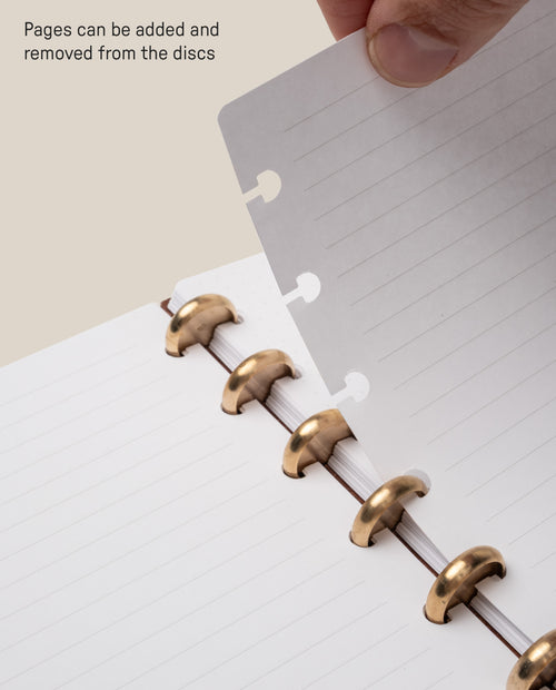 A hand adds or removes a lined notebook page with special holes from gold metal discs in the Heirloom Discbound Journal. Text reads, Pages can be added and removed from the discs—simply use the Discbound Journal Refill - Lined (Standard 6″ x 8.5″) pack to customize your journal.