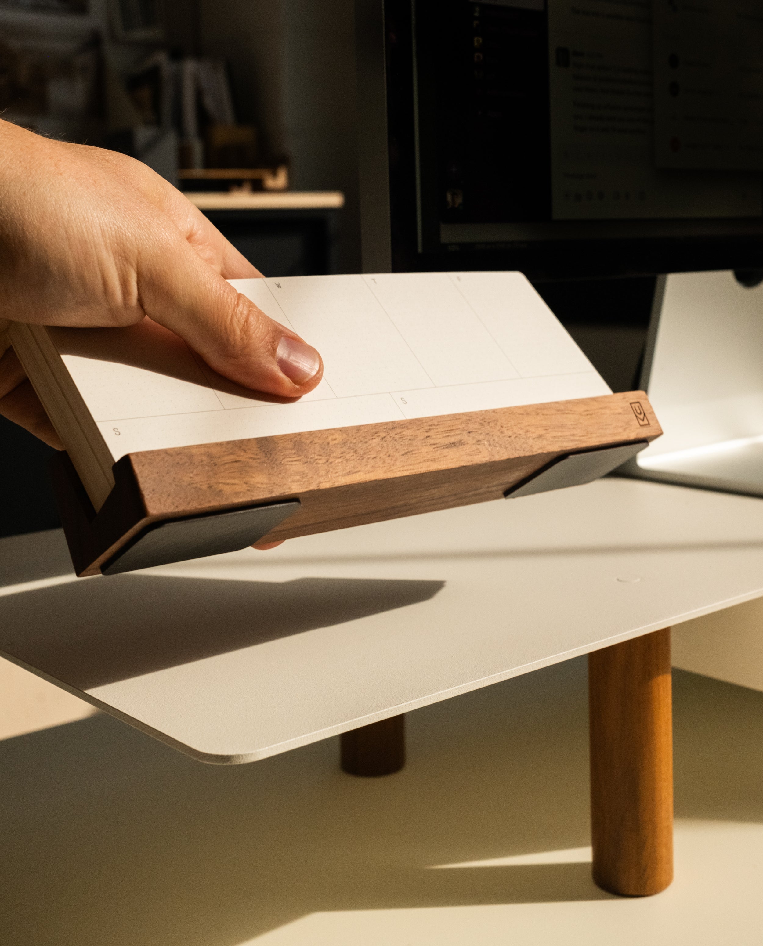 A hand holds a wooden desk calendar with blank pages near a computer monitor, which is elevated on a Magnet Kit (Pen Tray and Analog Weekly) atop a white desk with wooden legs. Sunlight shines across the surface, highlighting the organization and style of the workspace.